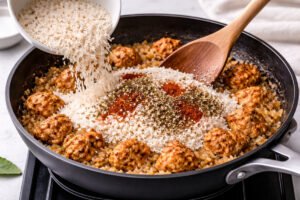 adding uncooked rice and spices to chicken and onion mixture in skillet